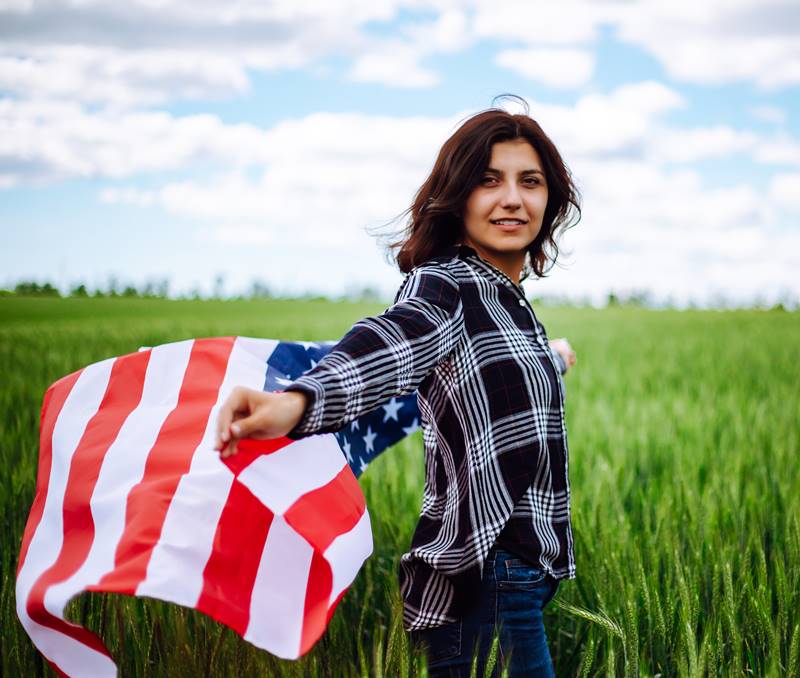 Young woman waves an american flag on the green wheat field. Patriotic holiday celebration. United States of America independence day, 4th of July concept